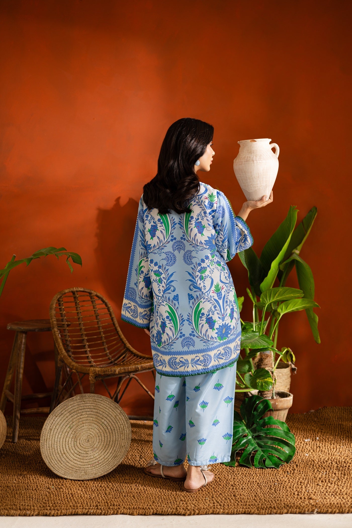 Woman holding a white vase against an orange wall with plants and furniture.